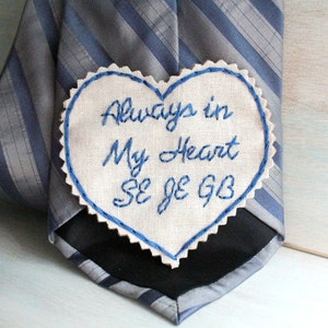 Può includere: Un cerotto di stoffa bianco a forma di cuore con ricamo blu che recita "Always in My Heart Se Je GB" attaccato a una cravatta a righe blu e grigie.