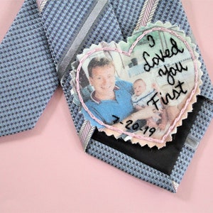 May include: A blue patterned tie with a pink heart-shaped patch sewn on. The patch has a photo of a man and a baby with the text "I loved you first" and the date "7-2019" embroidered on it.