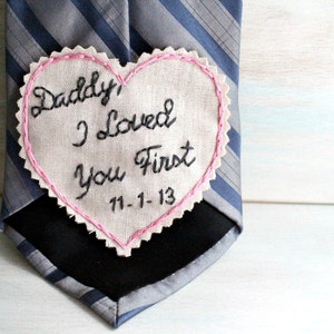 Könnte beinhalten: Ein herzförmiger Stoff-Patch mit rosa Nähten, der auf eine gestreifte Krawatte genäht ist. Der Patch ist mit dem Text "Daddy, I loved you first 11-1-13" bestickt.