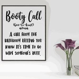 May include: A framed print with the words "Booty Call" and its definition in black text. The print is on a white surface, with a vase of purple flowers and a small potted plant nearby.