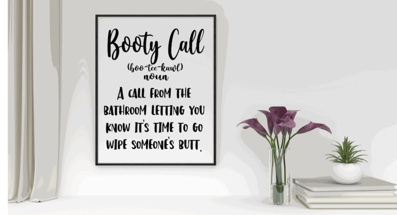 May include: A framed print with the words "Booty Call" and its definition in black text. The print is on a white surface, with a vase of purple flowers and a small potted plant nearby.