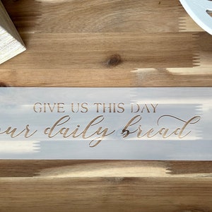 May include: A white stencil with the text "Give us this day our daily bread" in gold lettering. The stencil is on a wooden surface.