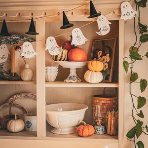 Könnte beinhalten: Ein Schrank mit Halloween-Dekoration. Eine Girlande aus Hexenhüten und Geister-Ausschnitten hängt oben. Auf den Regalen sind Kürbisse in verschiedenen Größen und Farben ausgestellt, zusammen mit dekorativen Tellern, Schalen und einem gerahmten Bild.