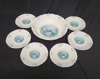 May include: A set of seven white porcelain plates with gold trim and a blue and white swan design. The set includes a large serving bowl and six smaller plates.