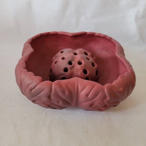 May include: A deep red ceramic flower frog with a round, perforated center and a leaf-shaped outer rim.