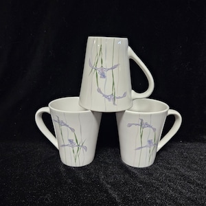 Puede incluir: Tres tazas de cerámica blanca con un diseño floral que presenta iris púrpuras y tallos verdes. Las tazas están apiladas una encima de la otra.