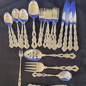 Könnte beinhalten: Ein Besteckset mit verzierten Griffen. Das Set enthält Löffel, Gabeln, Messer, einen Servierlöffel, eine Serviergabel und ein Buttermesser. Das Besteck ist aus poliertem Edelstahl.