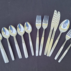 Puede incluir: Un conjunto de cubiertos de acero inoxidable, que incluye cucharas, tenedores y cuchillos, dispuestos sobre una superficie oscura. Los cubiertos presentan un patrón floral decorativo en los mangos. Las cucharas, tenedores y cuchillos son reflectantes.