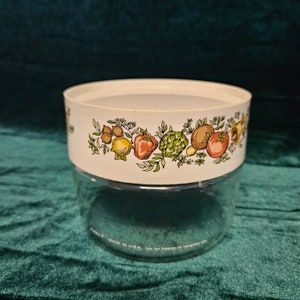 Puede incluir: Un tarro de vidrio transparente y blanco con una tapa blanca. La tapa tiene un borde colorido con un patrón de frutas, verduras y hierbas. El tarro tiene el texto "Made in U.S.A. Not for freezer or microwave use" en la parte inferior.