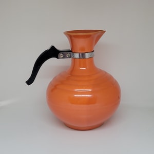 May include: Orange ceramic jug with a black plastic handle and a silver metal band around the neck.