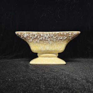 May include: A yellow ceramic planter with a brown and white speckled glaze. The planter has a wide, oval-shaped top and a pedestal base.