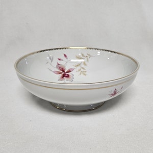 Könnte beinhalten: Eine weiße Porzellanschale mit goldfarbenem Rand und einem Blumendesign mit rosa und violetten Blüten. Die Schale hat einen Fuß.