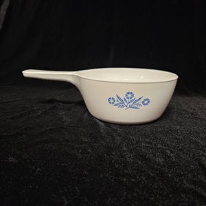 Puede incluir: Un cuenco blanco con asa y diseño floral azul. El cuenco tiene forma redondeada y un asa larga que se extiende desde un lado. El diseño floral se repite en el lateral del cuenco.