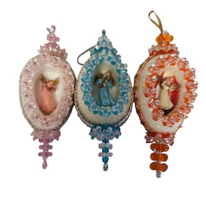 Puede incluir: Tres adornos navideños vintage. Cada adorno presenta una pequeña figura de ángel dentro de un marco ovalado. Los marcos están decorados con cuentas de colores rosa, azul y naranja. Los adornos tienen lazos dorados para colgar. Miden unos 7 cm de largo.