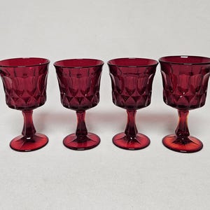 May include: Four ruby red wine glasses with diamond-patterned bowls and stemmed bases. The glasses are evenly spaced and set against a plain white background. The glasses are likely vintage or antique.