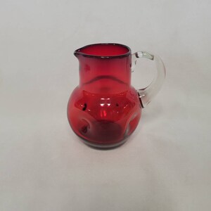May include: A small, red glass pitcher with a clear glass handle. The pitcher has a round body with a textured surface.