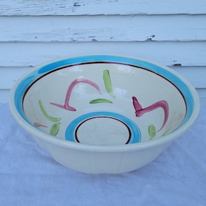 Puede incluir: Un tazón grande de cerámica blanca con un diseño pintado a mano en azul, marrón, verde y rosa. El tazón tiene un borde ancho y un fondo redondeado.