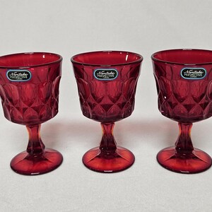 May include: Three ruby red stemmed glasses with diamond-cut patterns. Each glass has a round base and a short stem. The glasses have a label that reads "Noritake Perspective".