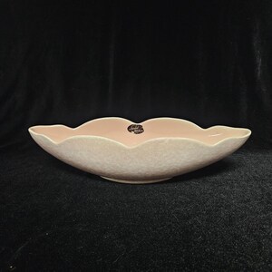 May include: A light pink, scalloped-edge ceramic bowl with a speckled exterior. The bowl has a wide, shallow design and a smooth interior. A small, circular label is visible inside the bowl. The bowl is likely for serving food or as a decorative piece.