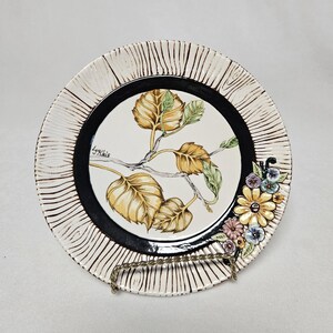 Peut inclure: Une assiette en céramique blanche avec un bord noir et un motif peint de feuilles et de fleurs. L'assiette a un bord texturé et est sur un support.