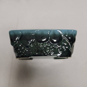 May include: A green and blue glazed ceramic planter with a grapevine design. The planter has a rectangular shape with rounded edges and four small feet.