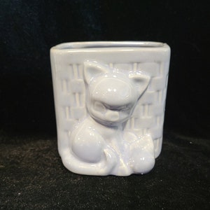 以下が含まれることがあります： 編まれたバスケットに座っている猫の形をした、薄い青色のセラミック製のプランター。プランターは滑らかで光沢のある仕上げです。