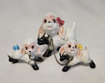 May include: Three porcelain figurines of lambs, each with black legs and a white body. The largest lamb has a pink face and a red bow, while the two smaller lambs have blue and yellow bows. The lambs are connected by a gold chain.