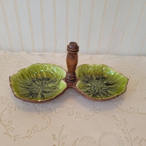 May include: A green ceramic serving dish with two sections and a wooden handle. The dish is shaped like leaves and has a mottled green glaze.