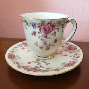 May include: A white teacup and saucer set with a floral pattern in shades of pink, yellow, and blue. The cup and saucer have a gold trim.