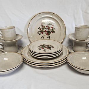 May include: A full beige stoneware dinner service featuring a strawberry design. The set includes plates, bowls, saucers, and cups. The plates have a scalloped edge and a brown trim. The strawberry design features red berries and green leaves.