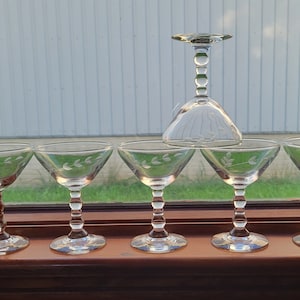 Peut inclure: Ensemble de six verres à cocktail en verre transparent à pied avec un délicat motif de feuilles gravées. Les verres sont disposés en rangée sur une surface en bois.