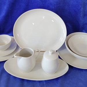 May include: A set of white ceramic dishes, including a large platter, a smaller oval platter, a bowl, a creamer, a sugar bowl, a cup, and a saucer.