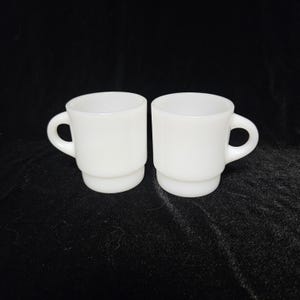 Pode incluir: Duas canecas de café brancas empilháveis com alças arredondadas. As canecas têm um design clássico e minimalista, perfeito para servir café ou chá. As canecas são feitas de um material sólido e opaco.
