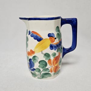 May include: A white ceramic pitcher with a blue handle and rim. The pitcher is decorated with a colorful bird and floral design in yellow, orange, blue, and green. The pitcher is approximately 8 inches tall.
