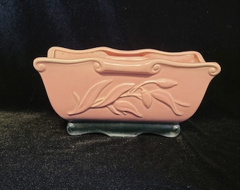 May include: A pink ceramic planter with a leafy design and a dark green base. The planter is shaped like a trough and has a scalloped edge.