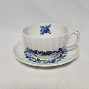 Puede incluir: Una taza de té y un platillo blancos con un patrón floral azul y verde. La taza tiene una superficie texturizada y un asa. El platillo tiene un borde festoneado.