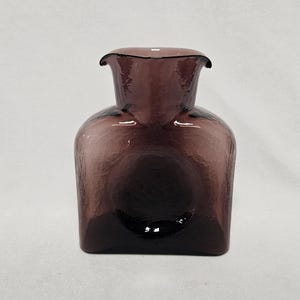 Puede incluir: Un jarrón de vidrio morado oscuro con una superficie texturizada. El jarrón tiene una base cuadrada y una parte superior redondeada con un borde acampanado. El vidrio es translúcido.