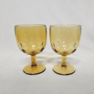 Pode incluir: Duas taças de vinho âmbar. Cada taça tem uma taça arredondada com um padrão texturizado e uma haste. Os óculos são feitos de vidro e estão em um fundo simples.