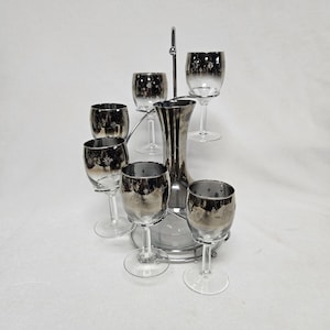 Pode incluir: Um conjunto de bar vintage prateado com um jarro e seis copos. Os copos e o jarro têm um efeito ombré prateado e um padrão de estrelas. O conjunto é exibido em um suporte de metal prateado.