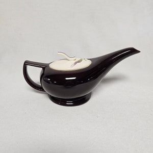 Könnte beinhalten: Eine dunkelbordeauxfarbene Teekanne aus Keramik in Form einer Genielampe mit einem weißen Deckel und einem Blumendesign. Die Teekanne hat einen Ausguss und einen Griff.