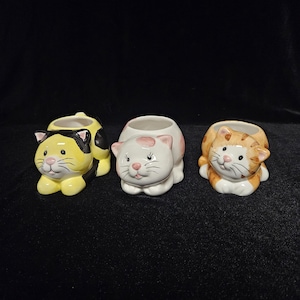 Puede incluir: Tres macetas de cerámica con forma de gato en amarillo, blanco y naranja. El gato amarillo tiene marcas negras, el gato blanco tiene manchas rosas y el gato naranja tiene rayas marrones. Cada maceta tiene una abertura redonda.