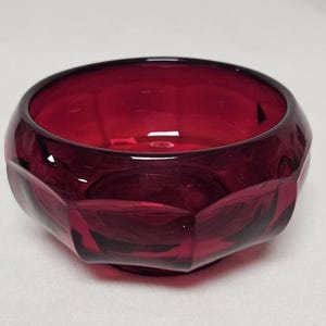 May include: A ruby red glass bowl with a faceted exterior. The bowl has a wide, open top and a rounded base. The glass is transparent, allowing light to pass through. The bowl is likely used for decorative purposes or serving.