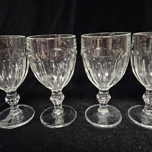 May include: Four clear wine glasses with a ridged design on the bowl. The glasses have a classic shape with a round base and a detailed stem. The background is black. Ideal for serving wine.