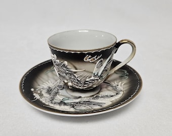 Könnte beinhalten: Ein schwarzes und weißes Tee-Set mit Drachenmotiv. Tasse und Untertasse sind mit einem silbernen und goldenen Drachen verziert.