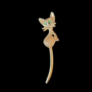 May include: A gold-toned brooch in the shape of a cat with green gem eyes. The cat's tail is curled and the brooch has a pin back.