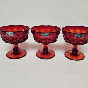 May include: Three ruby red glass dessert bowls with a diamond pattern and a stem. Each bowl has a round base and a label. The bowls are arranged in a row against a white background.