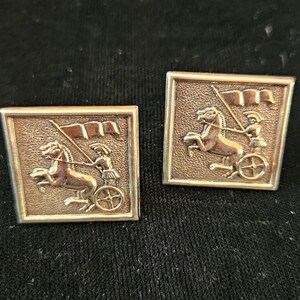 May include: Pair of vintage silver-toned cufflinks. Each cufflink features a raised relief of a chariot pulled by a horse, driven by a figure holding a flag, set within a square frame. The background is a dark, textured fabric.