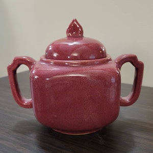 Könnte beinhalten: Eine quadratische, burgunderfarbene Teekanne aus Keramik mit Deckel und zwei Griffen. Die Teekanne hat eine glänzende Oberfläche und eine leicht strukturierte Oberfläche.