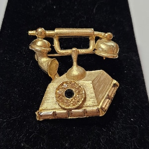 May include: A gold-tone rotary phone brooch with clear rhinestone accents. The phone has a black center and a gold-tone base.
