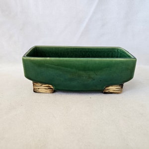 Pode incluir: Um vaso de cerâmica verde com quatro pés curtos pintados de ouro. O vaso é retangular e tem um acabamento liso e esmaltado.
